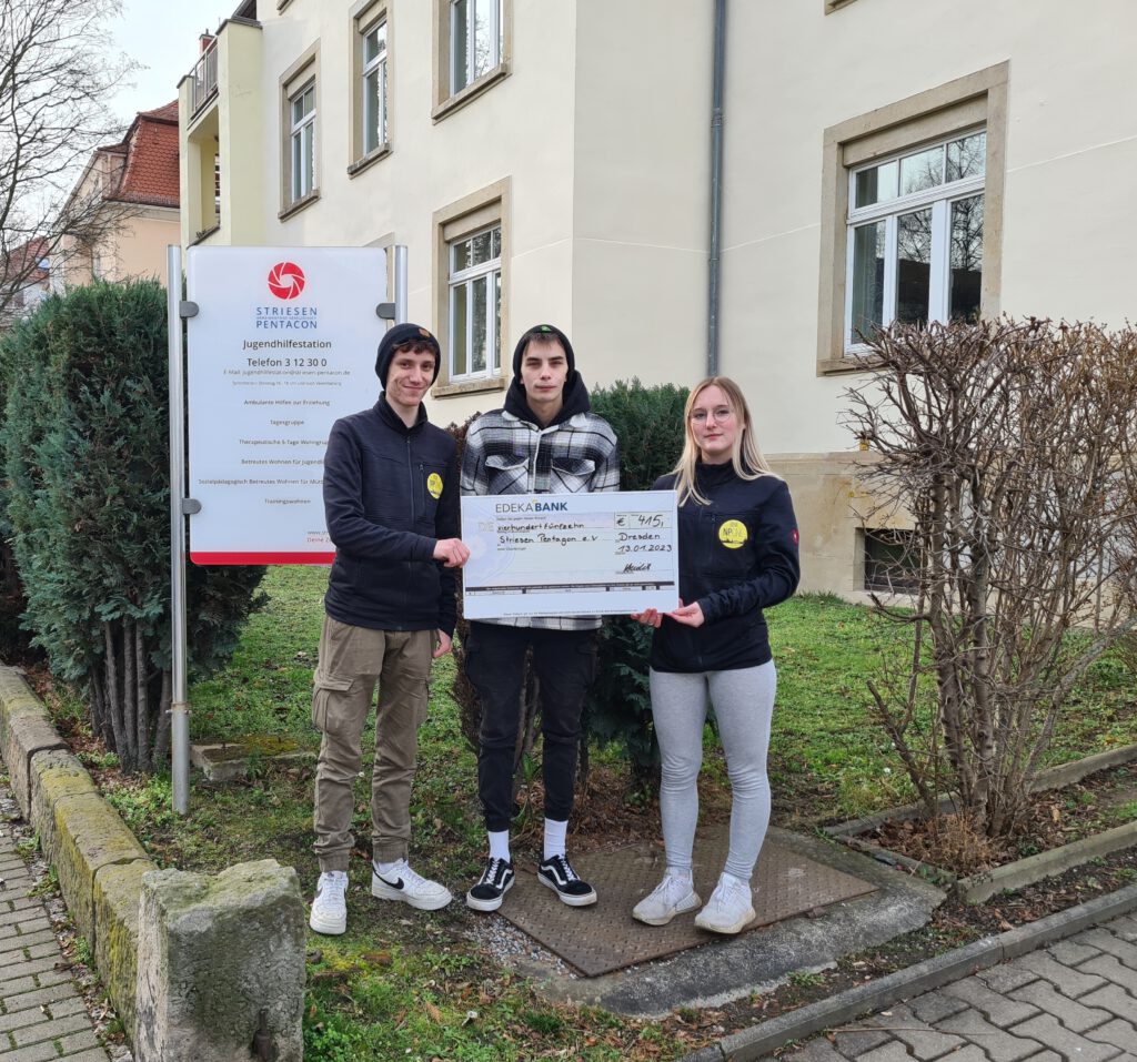 3 Personen halten einen Spendencheck vor der Einrichtung der Jugendhilfestation.